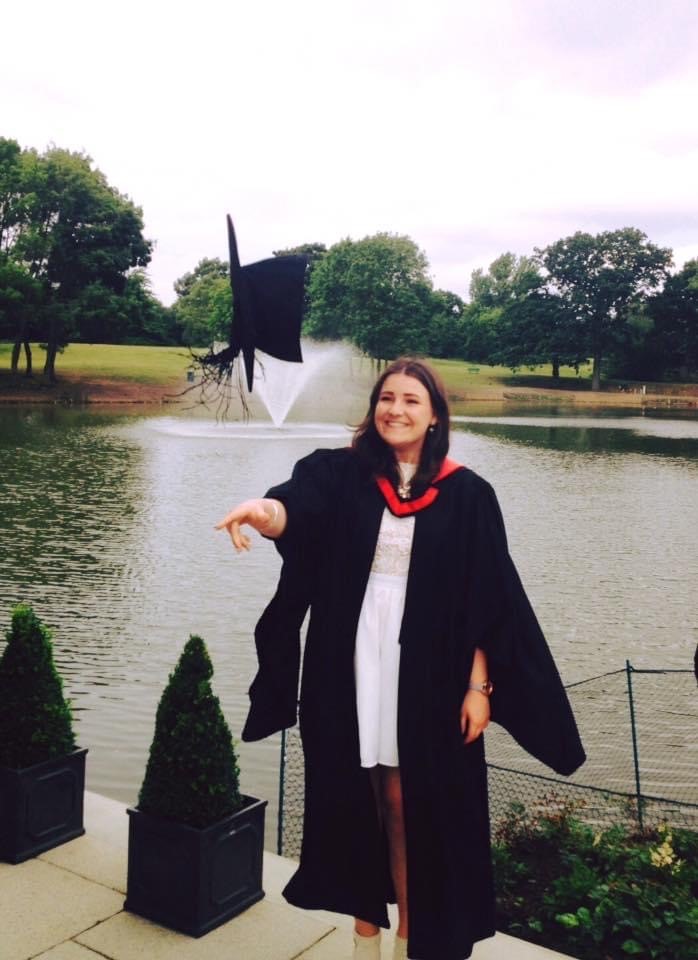 Annabel in her graduation robes in front of the lake on Colchester Campus, throwing her cap in the air.