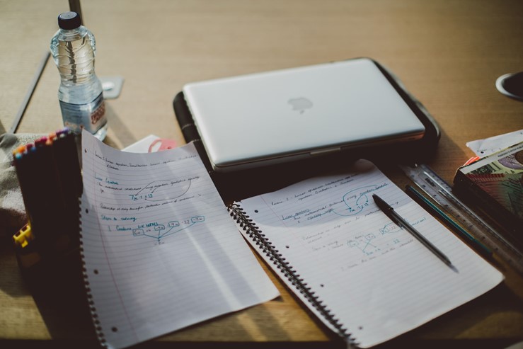 A photo of a desk with a laptop, writing notepaper, pens and other items on top.