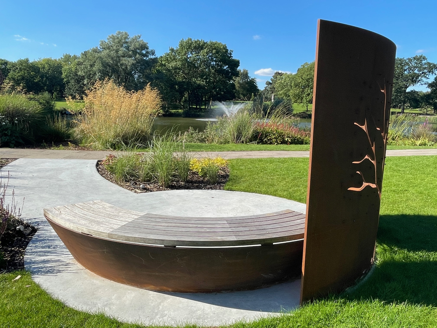 Our Tree of Life memorial in the sunshine. A curved bench and tree cut from a metal wall by the lake.