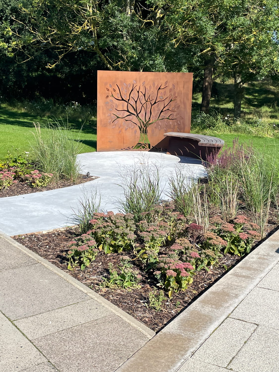 Our Tree of Life memorial in the sunshine. A curved bench and tree cut from a metal wall by the lake.