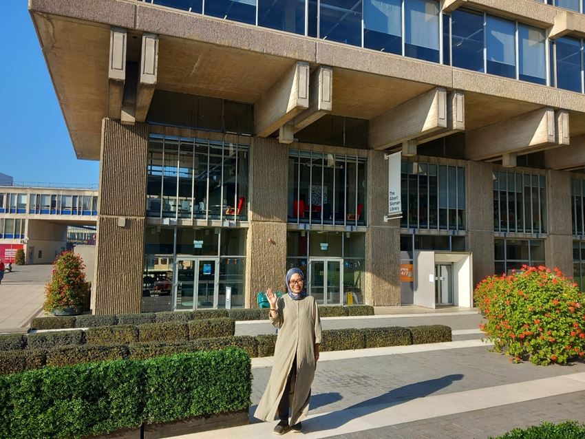 Aisyah Shamshun waving and standing outside the Albert Sloman Library.