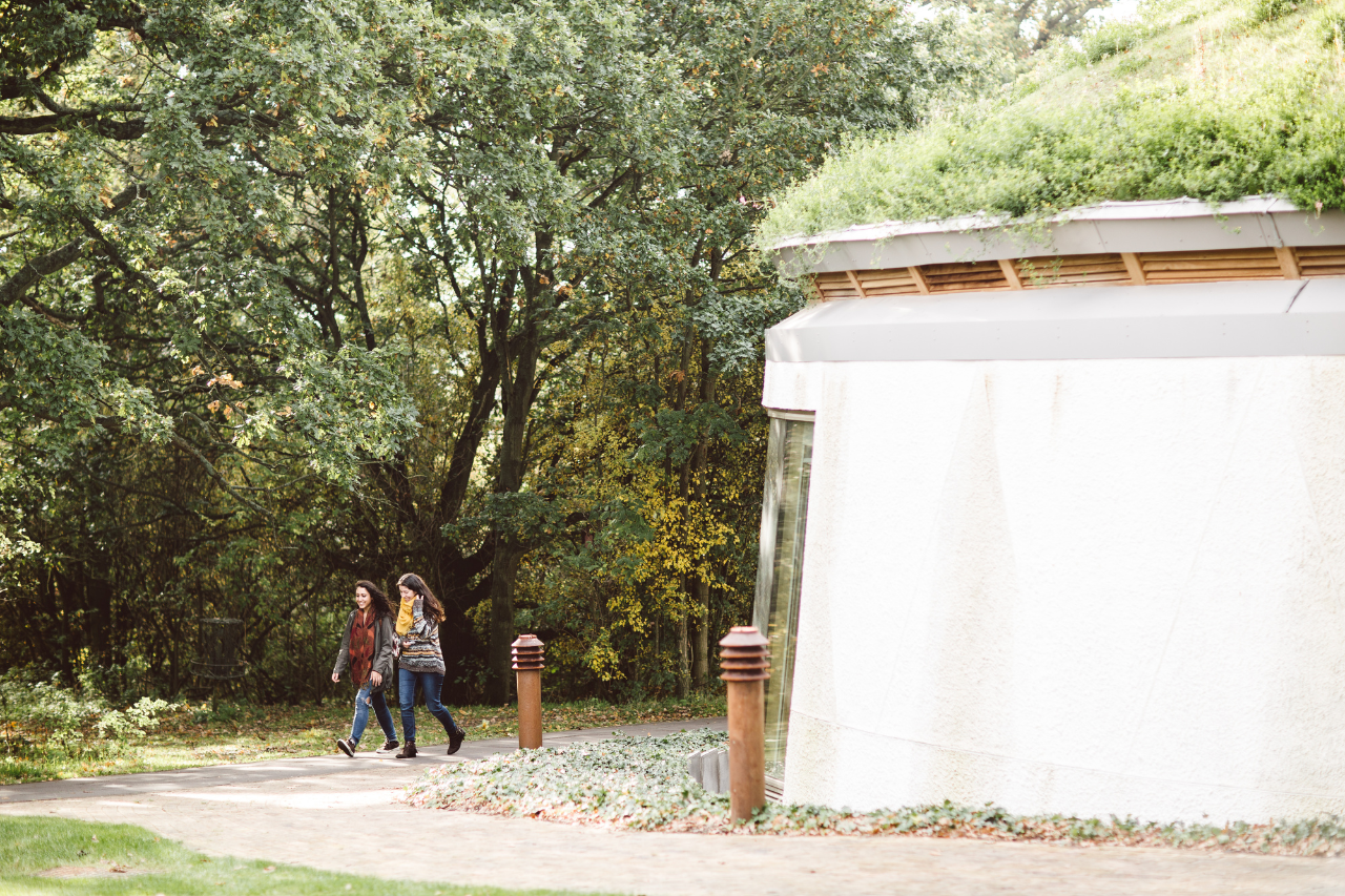 Two women walking outside of the EBS building in front of trees and a building with a grass roof