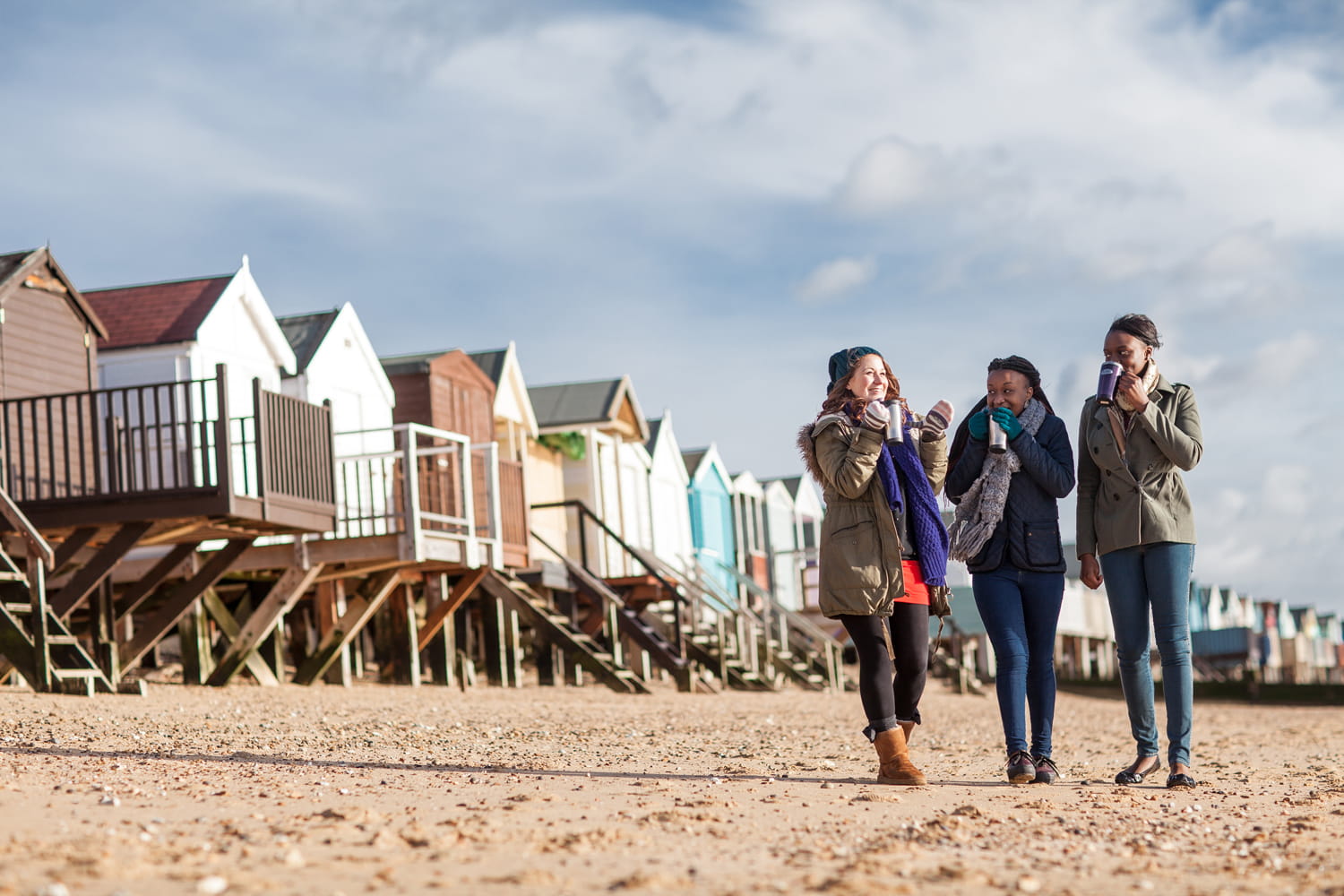 Three people walk along a sandy beach past beach huts.