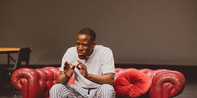 Jubril Agbaloo performs his piece She's Very Gorgeous To Me on the Lakeside Theatre main stage. Jubril is a young black man, wearing a pink tee shirt and pyjama bottoms and it sitting on a red leather sofa.