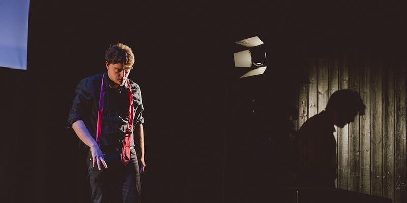 a young white man wearing a black shirt stands on the Lakeside Theatre main stage and the shot has captured his shadow against the black wall in dramatic fashion
