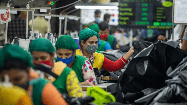 A row of women sitting at sewing machines in a factory, with lots of black fabric on the right hand side.