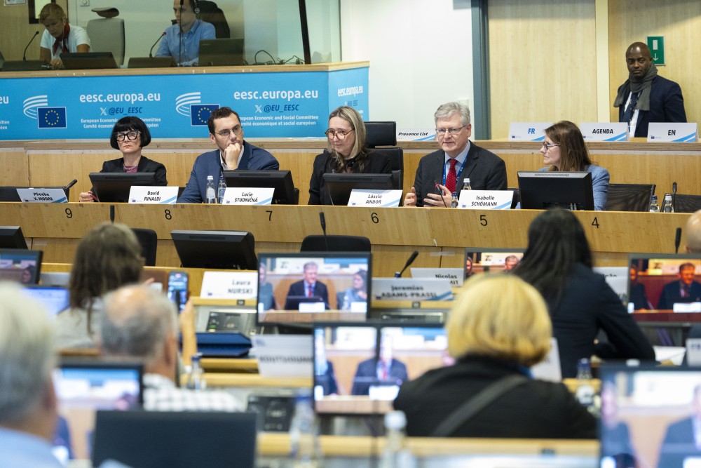 Five people sitting on a panel at a conference. In the foreground are people sitting at desks with screens in front of them.
