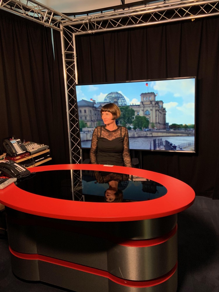 Professor Phoebe Moore sitting at a black table with red around the edge. A large TV screen is behind her.