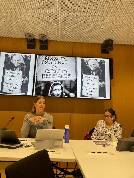 Two people sitting at a desk with a screen behind them. The person on the left is giving a talk.