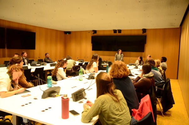 People sitting around tables set up in a large "U" shape. At the top someone is standing and addressing the room.