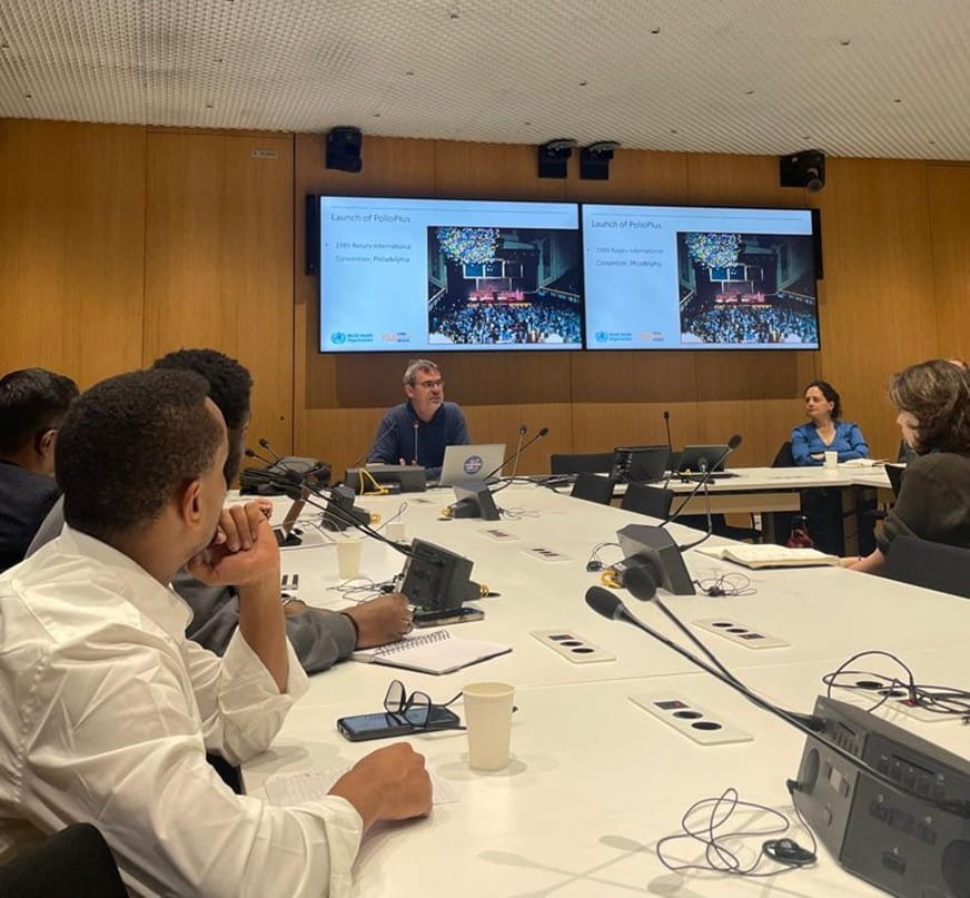 People sitting at a desk looking towards a speaker at the front of the room. Behind the speaker are two large screens with presentations slides on "Launch of PolioPlus".