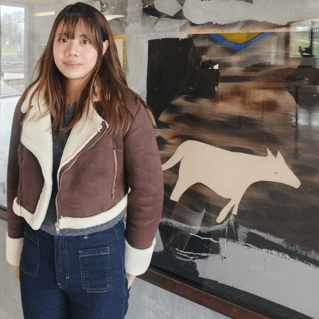 Joyce swearing a brown jacket and blue jeans, stood in front of a large piece of art.