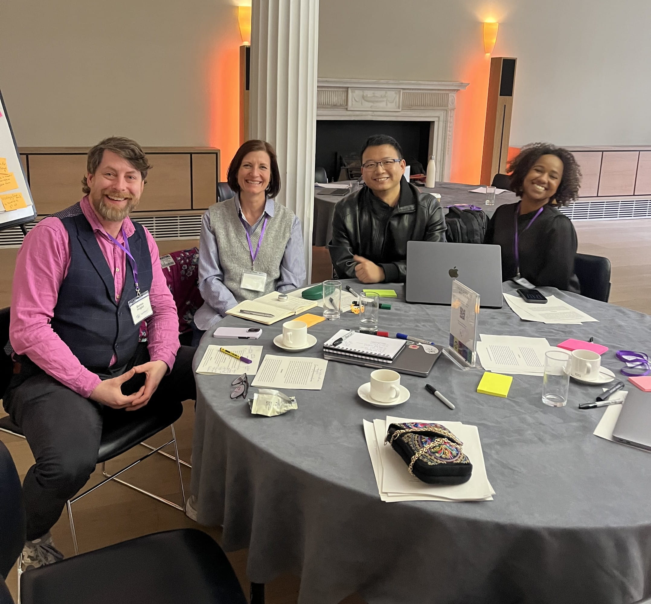 Four people sitting at a round table covered in papers, cups, glasses and a laptop.