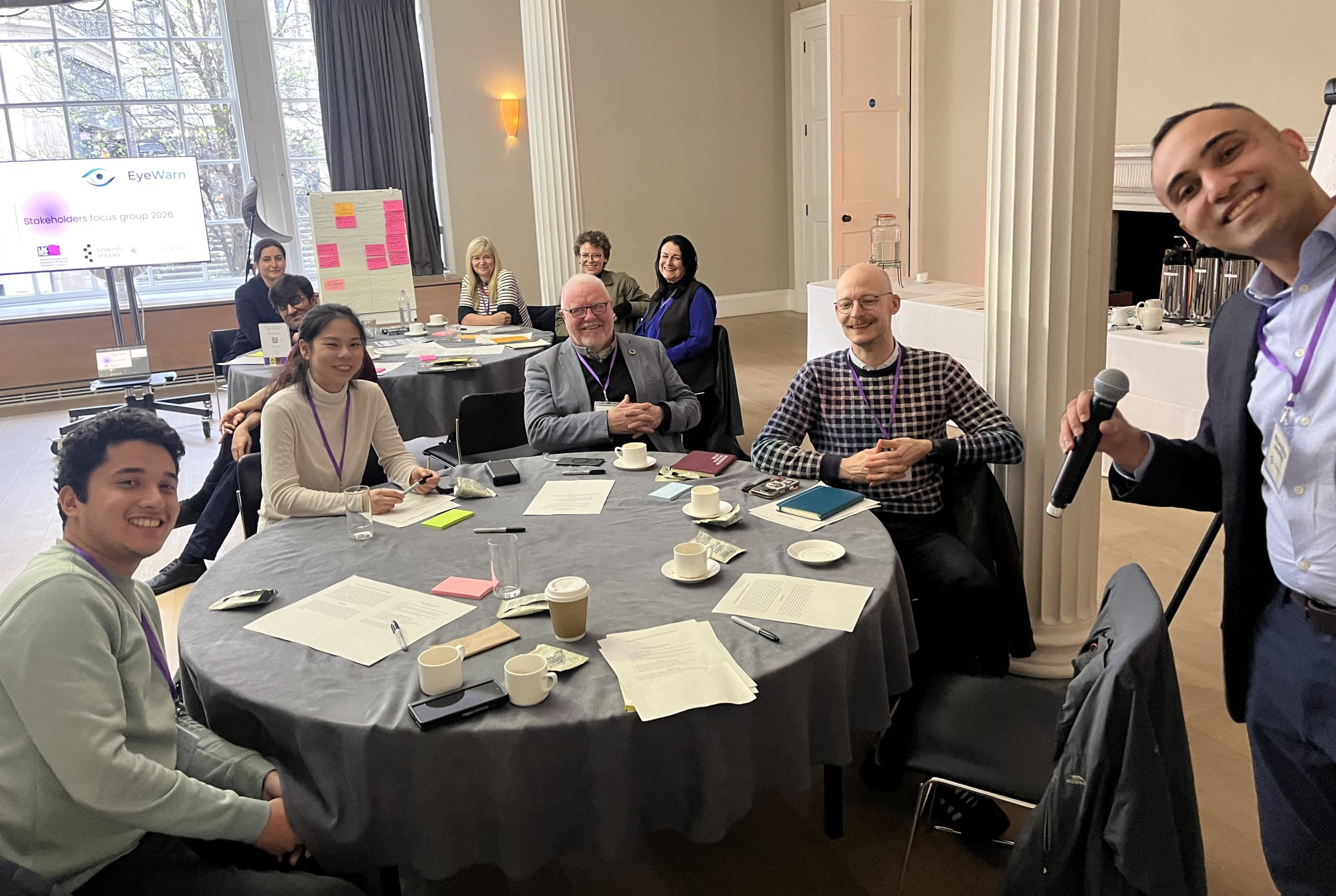 Two round tables with four people sitting at each one. The tables have papers and cups on them. A man holding a microphone is standing to the right smiling at the camera.