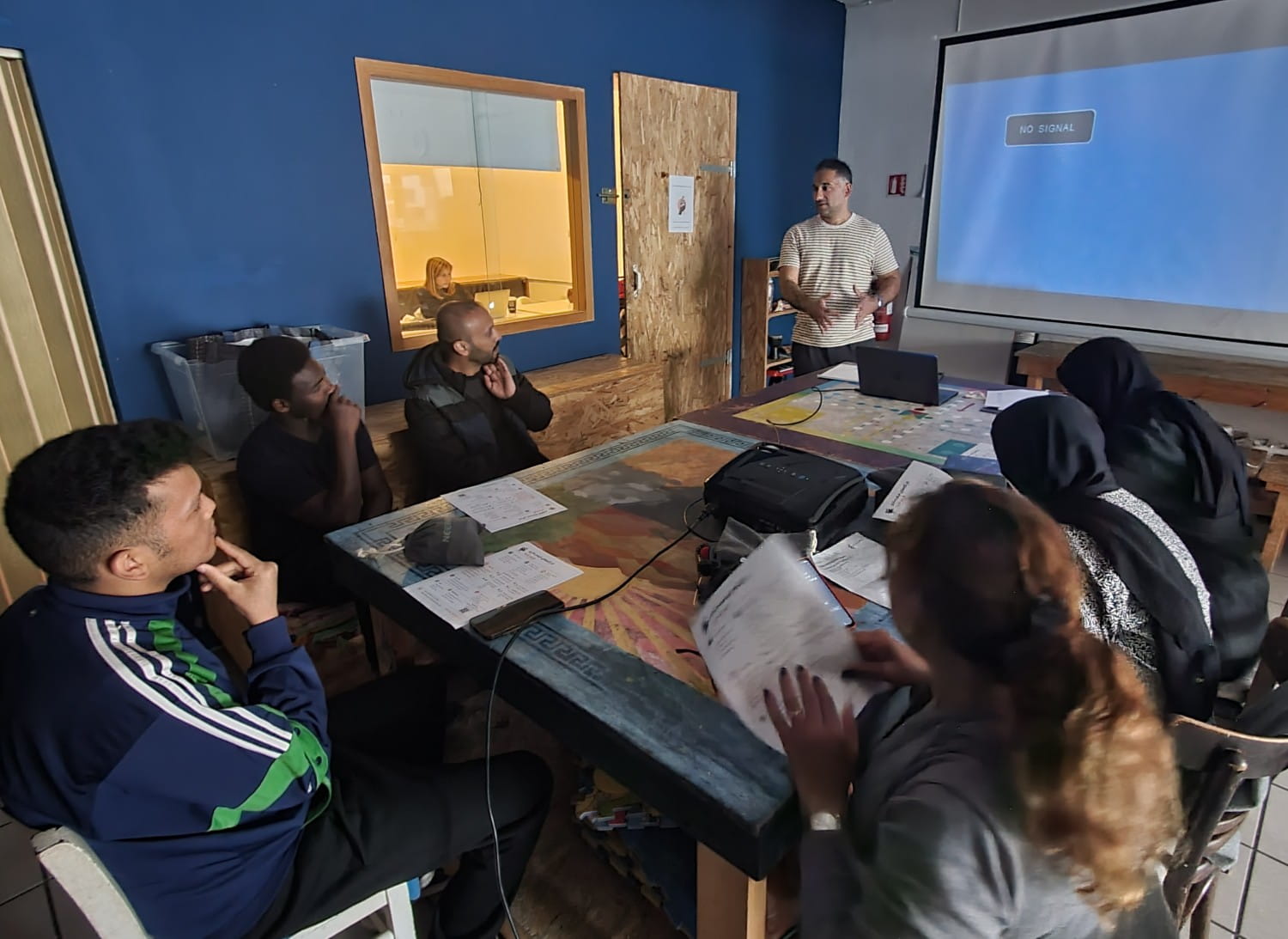 Six people sitting around a table, with Dr Tasos Giapoutzis standing at the front. The screen behind him is blank.