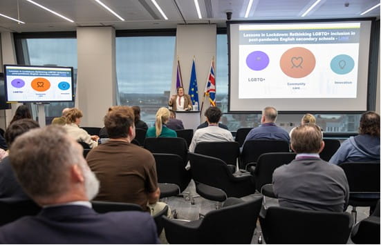 EJ Caris-Hamer standing a podium delivering a talk to a room of people. There are screens showing their slides on the left and right.