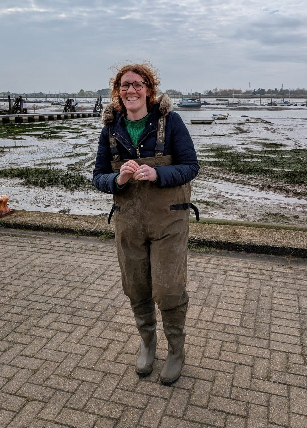Katy Wheeler wearing waterproof clothing, standing on a harbourside with water and sandy mud visible behind her.