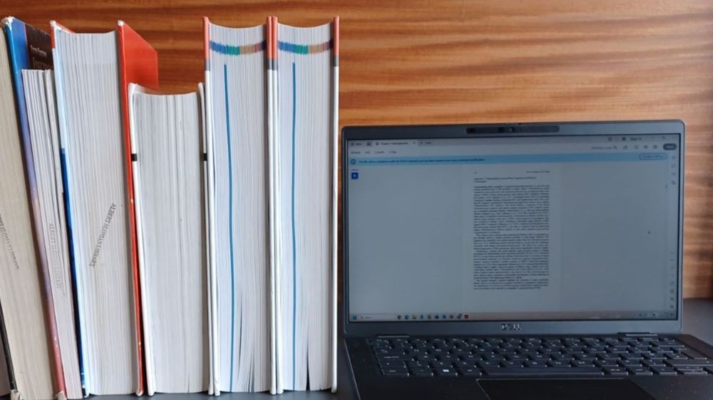 A row of six upright books, page edges to the front, shown next to a laptop with an open access digital text on the screen