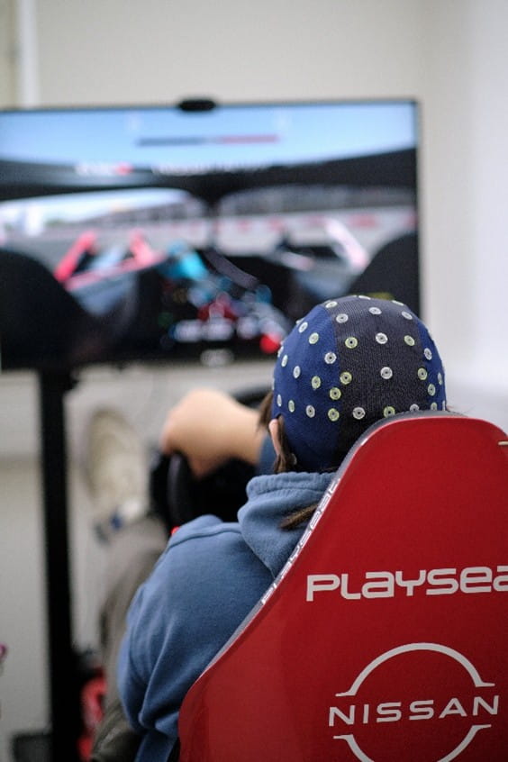 A person with their back to the camera, wearing a sensor cap sitting in a red gaming chair, playing a computer game with a steering wheel.