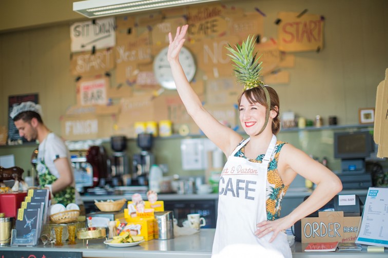 Photo of a female waitress waving her hand. She is wearing an apron which says CAFE and has a ornamental pipeapple on her head. In the background is the Lakeside Theatre Cafe.