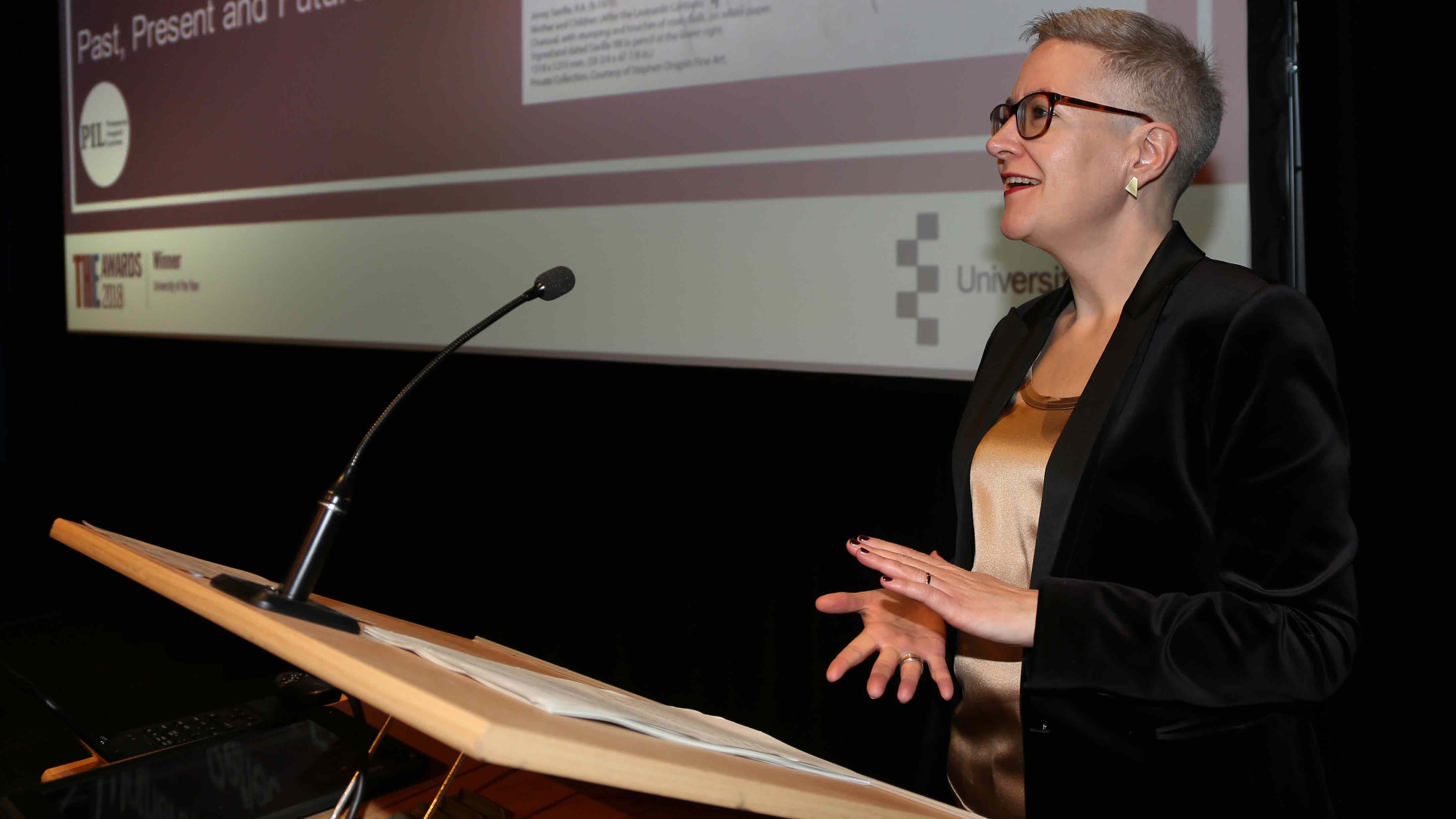 Professor Tracey Loughran, seen delivering a lecture, wearing a black jacket and dark-rimmed glasses