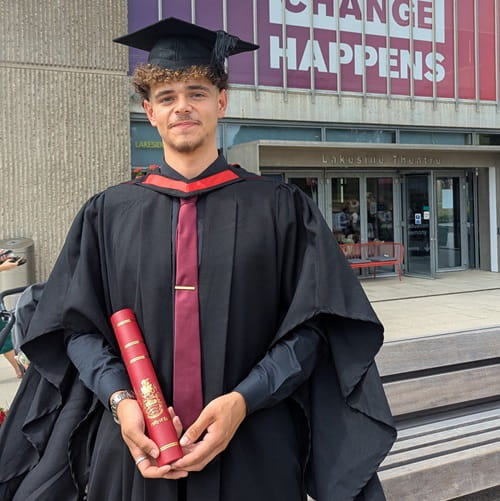 Football scholar Cameron Button dressed in graduation gown and mortarboard square shaped hat holding his certificate on graduation day at the University of Essex