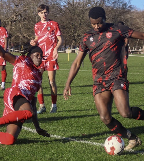 University of Essex football scholar Derrik Berko mid game on an outdoor grass pitch on the ball about to be tackled by an opponent wearing black and red Essex Blades kit