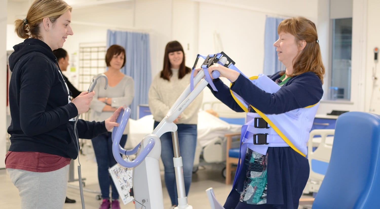 Erika Kerry from the School of Health and Social Care is standing on the left holding the control for an electronic hoist in her right hand. A mock-patient is on the right, with the hoist straps around her waist and under her arms. She is being lowered in to a blue chair. Two prospective students are standing in the background watching.