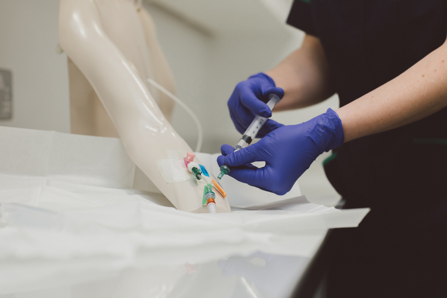 A person wearing purple gloves, their head out of shot, is practising using an intravenous injection on a plastic arm.