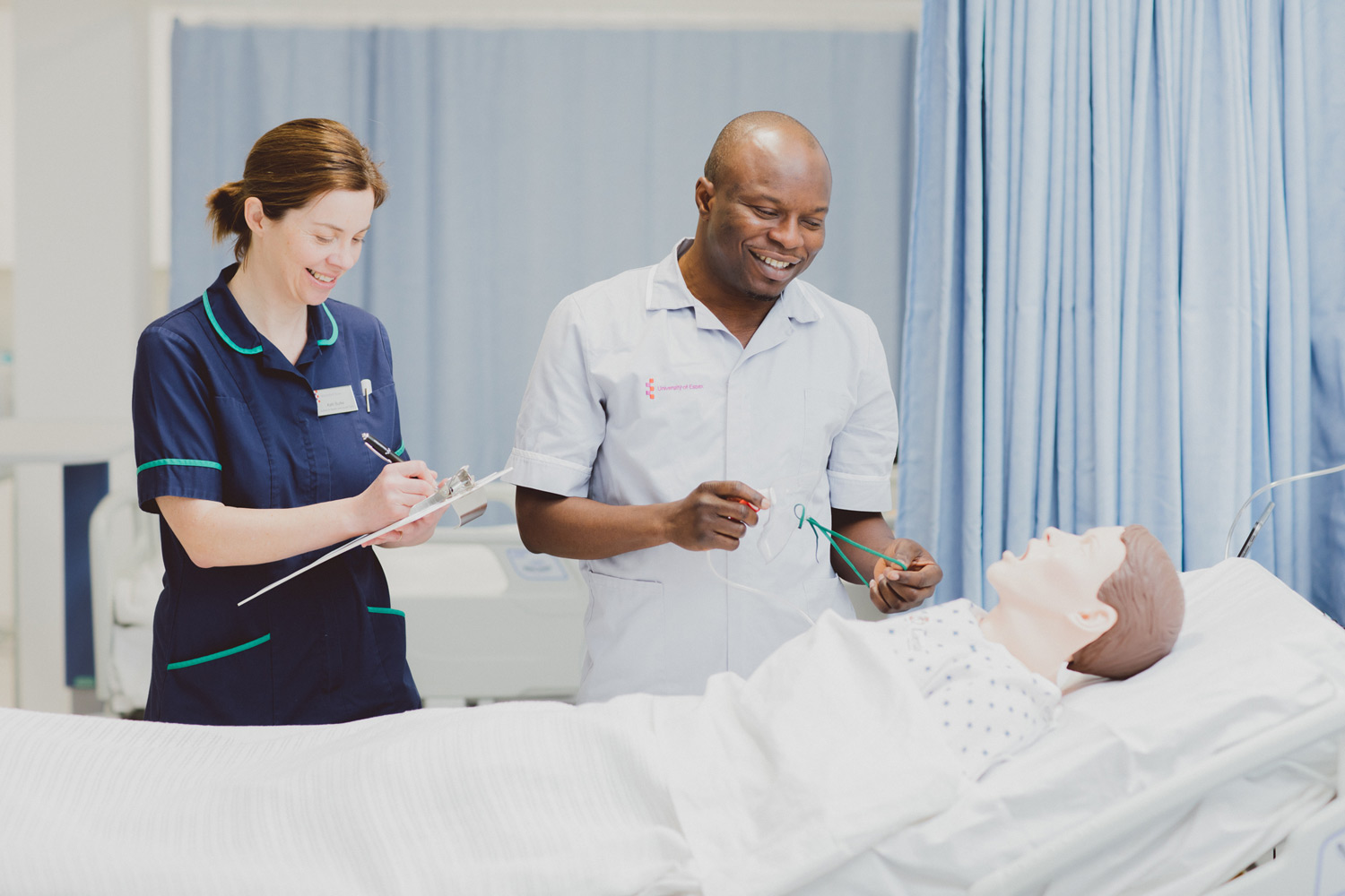 A nursing student in uniform, holding an oxygen mask, is standing next to a bed with a Sim person. A lecturer in a nurse uniform is standing to the left, writing something on a clipboard.