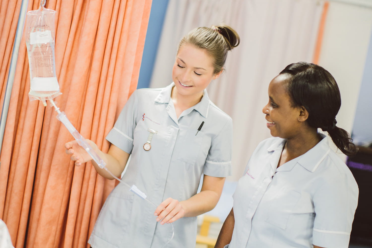 Two nursing students in uniform standing in a mock ward. One is holding a tube from an IV bag, showing it to the student next to her.