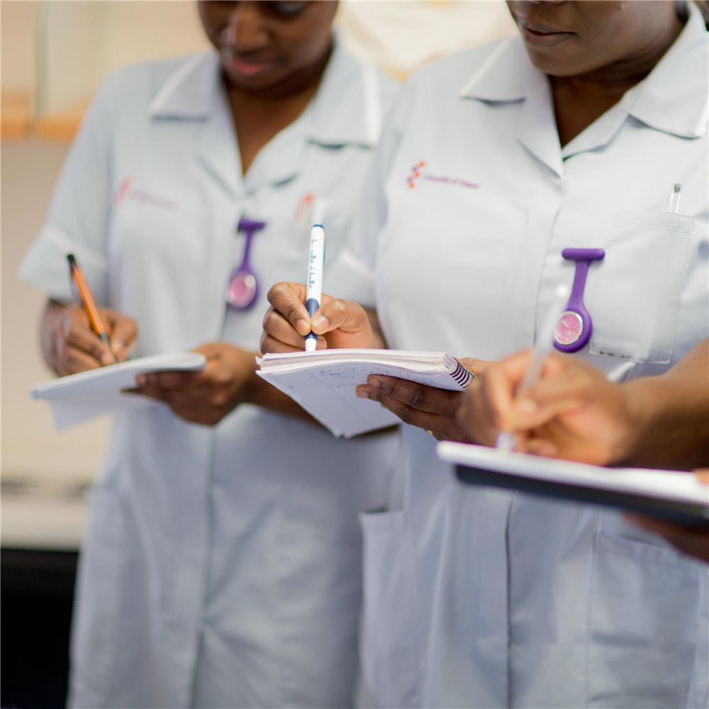Two students in nursing uniforms, and a third mostly out of shot on the right, standing and writing notes on some paper in their hands.