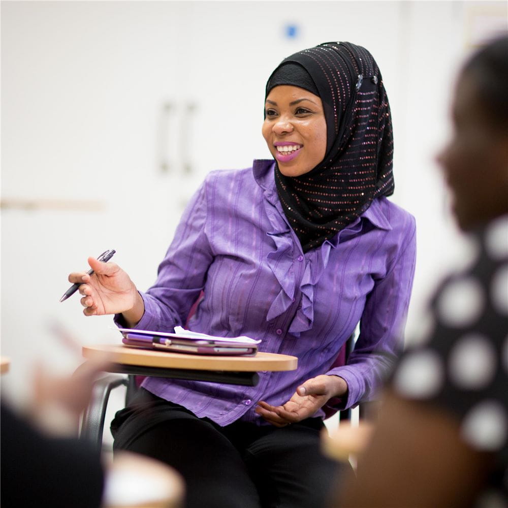 A student in a headscarf sitting in a seminar with a notebook in front of her, gesturing slightly with a pen in her right hand. A blurred second student is in the foreground to the right side of the photo.