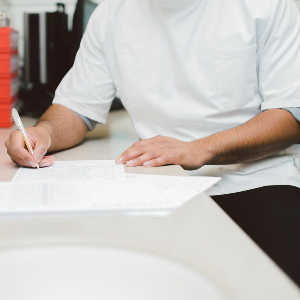 Photo of a person, with their head out of shot, sitting alongside a desk surface, writing on some pieces of paper on the desk.