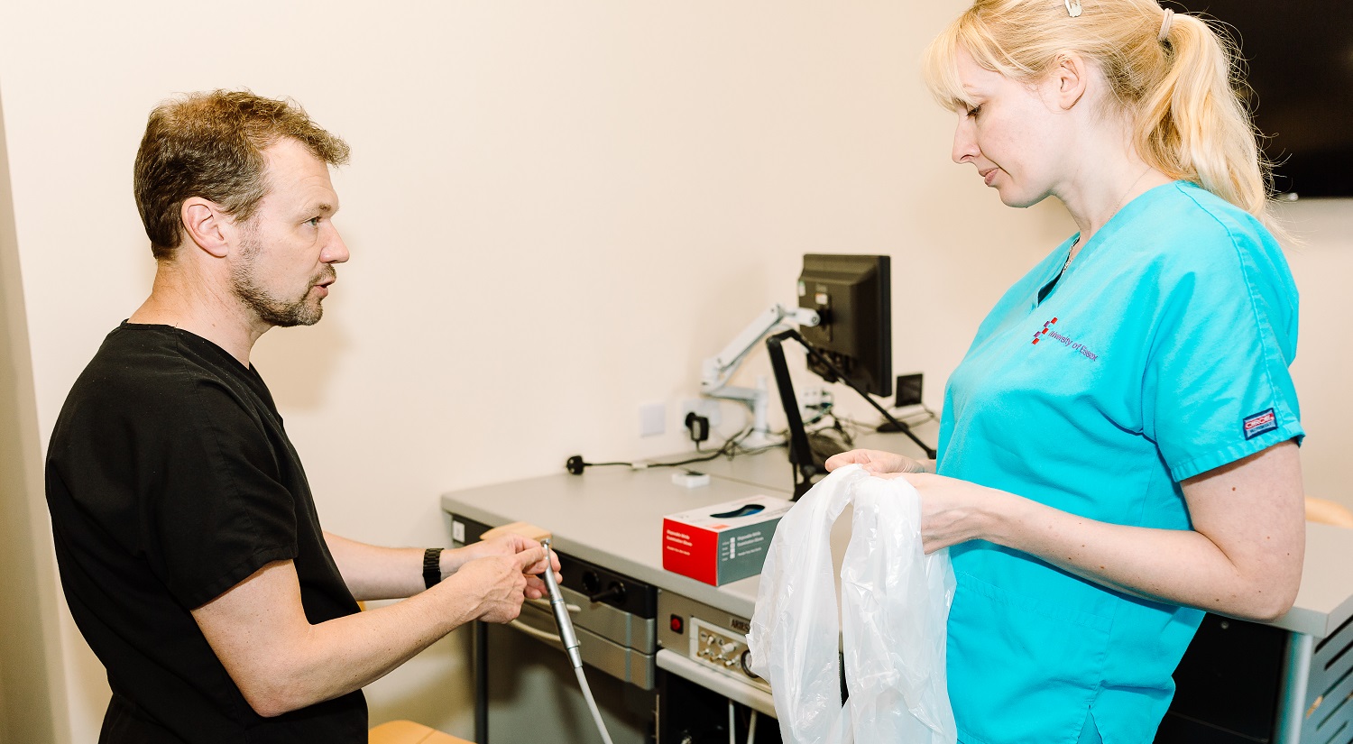 Nick Barker is standing on the left holding a dental drill. He is pointing at a piece of the drill and talking to an Oral Health Sciences student, who is standing to the right holding a plastic apron.