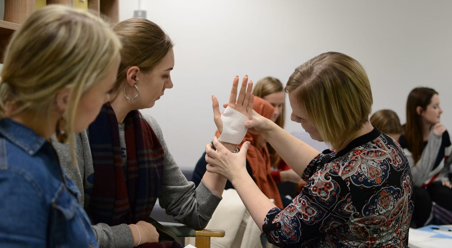 On the right, with her back to the camera, is Selena Hammond from the School of Health and Social Care. On the left are two students. The student in the middle has her elbow resting on the arm of her chair, her hand pointing up. Selena is holding a piece of sterile bandage against the hand, as part of a demonstration on how to make a splint. Several other students are sitting in the background.