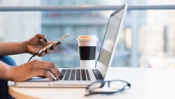 A pair of hands with black nail polish, one resting on the keyboard of a laptop on a desk and the other holding a phone with their thumb poised over the screen.