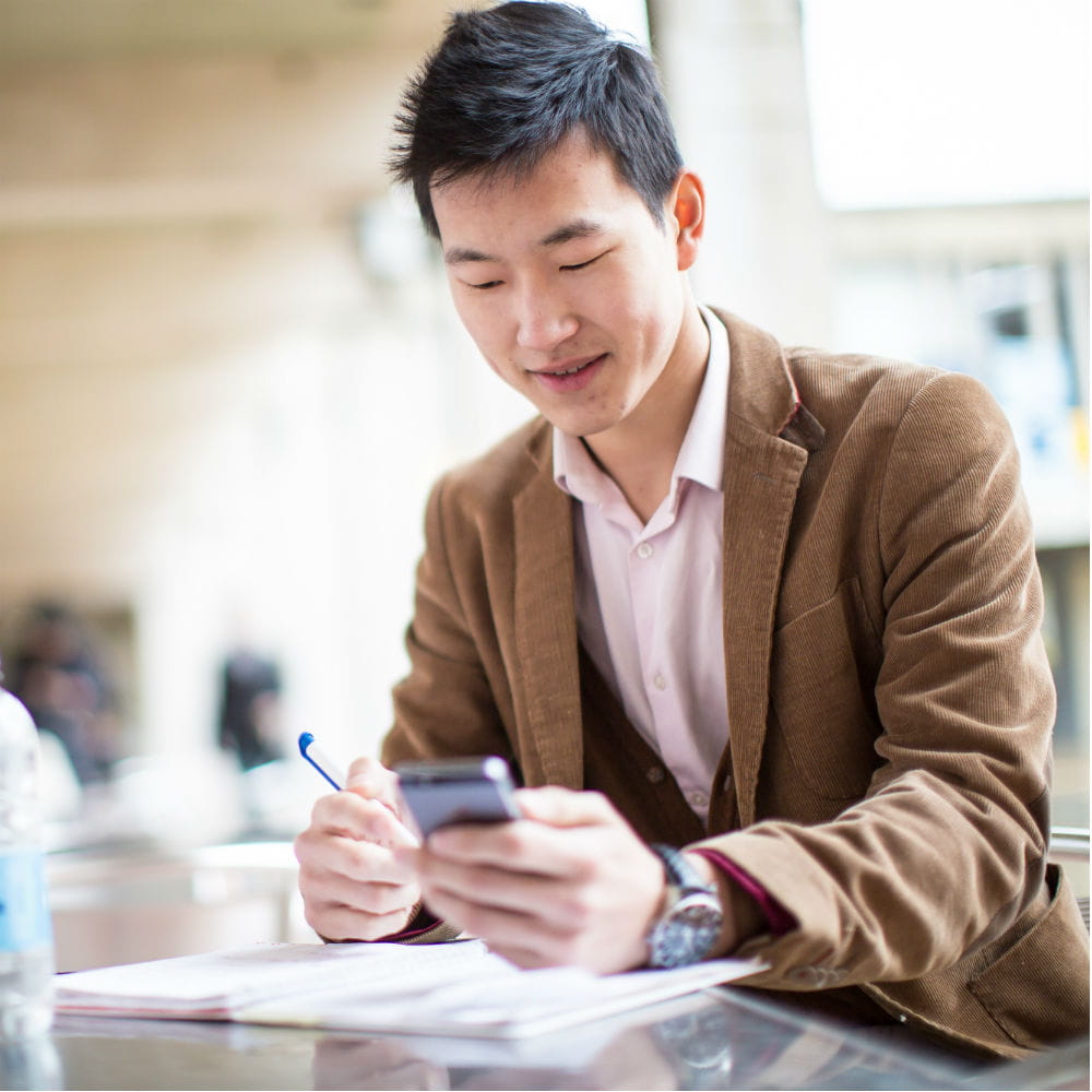 A man wearing a tan jacket sitting at a table with papers in front of him, looking at a phone in his hand.