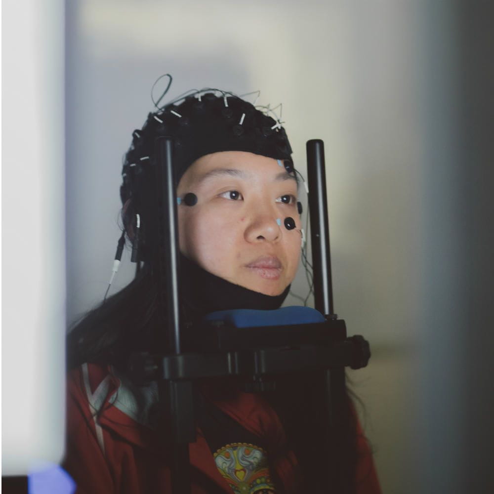 A student wearing a black EEG cap, resting her chin on a frame, with sensors attached to her face.