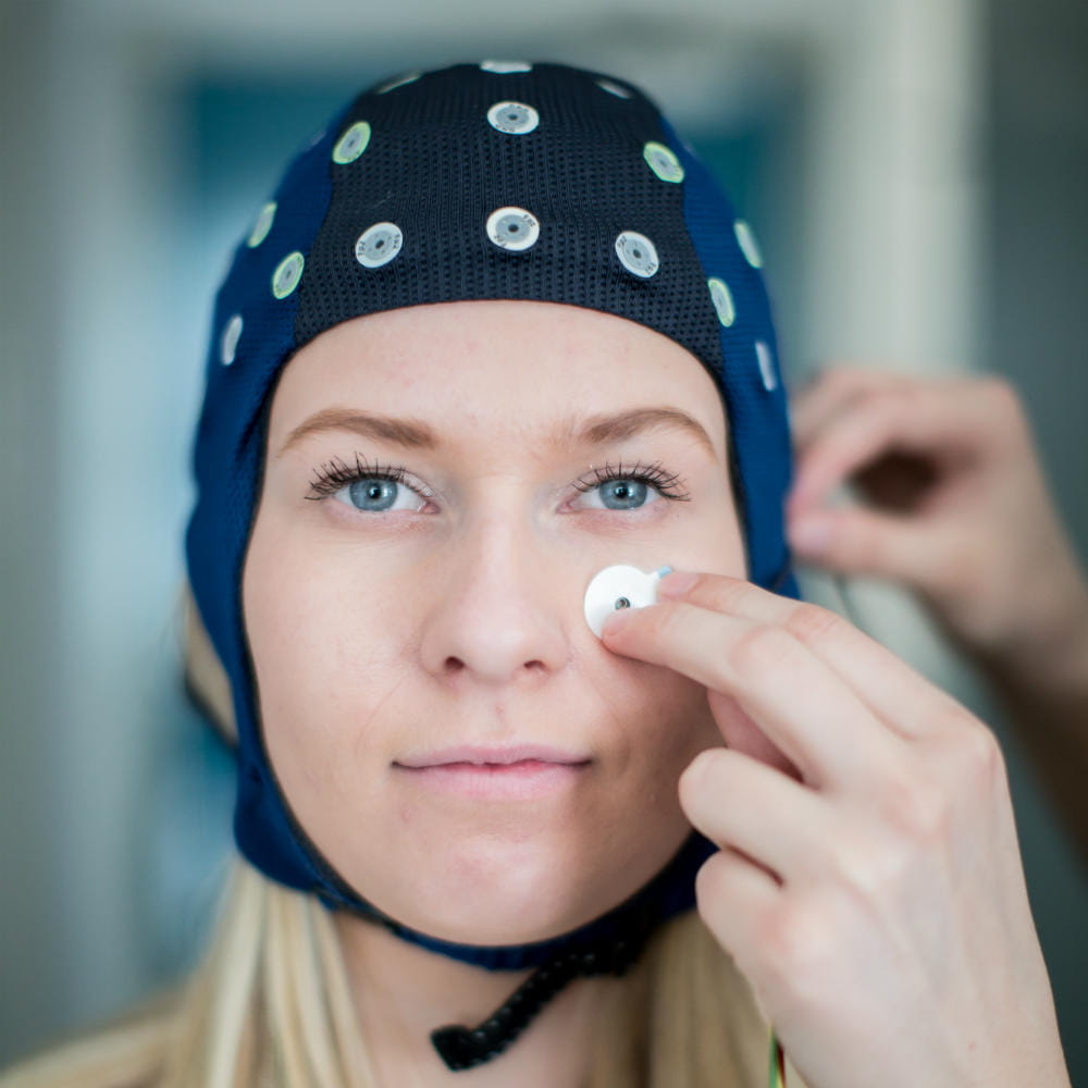 A woman with blonde hair wearing a blue EEG cap, with someone to the right attaching a small sensor to her cheek.