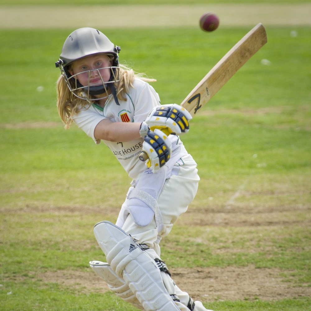 A young woman wearing cricket whites and a helmet, holding a cricket bat that she has just swung to hit a cricket ball.