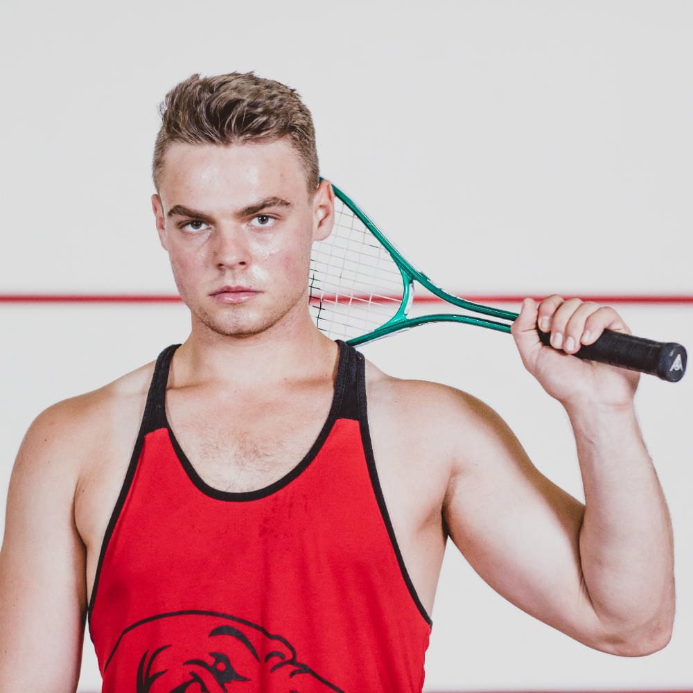A young man wearing a red sports vest and holding a tennis racquet up behind his head.