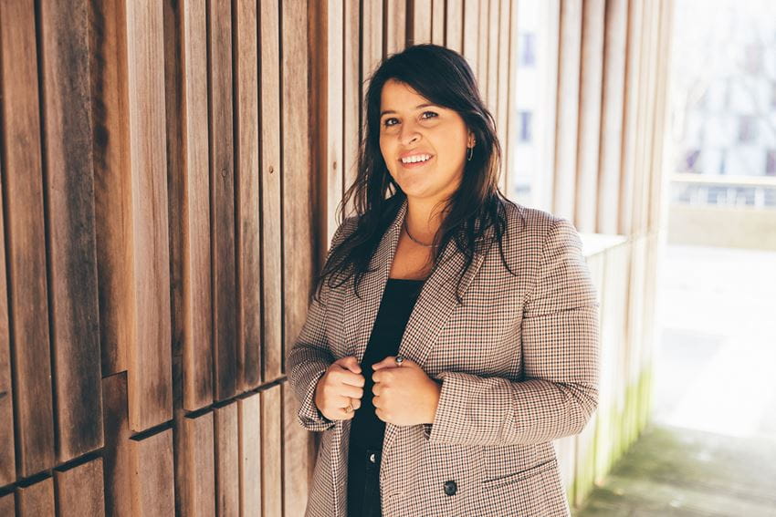 Mariana Zamudio Guajardo, Essex MBA student 2021-22, wears her mid-length dark hair down and smiles while wearing a grey suit jacket. She is standing outside the Essex Business School building and posing against a backdrop of one of the wooden structural features. She is smiling and looks very professional.