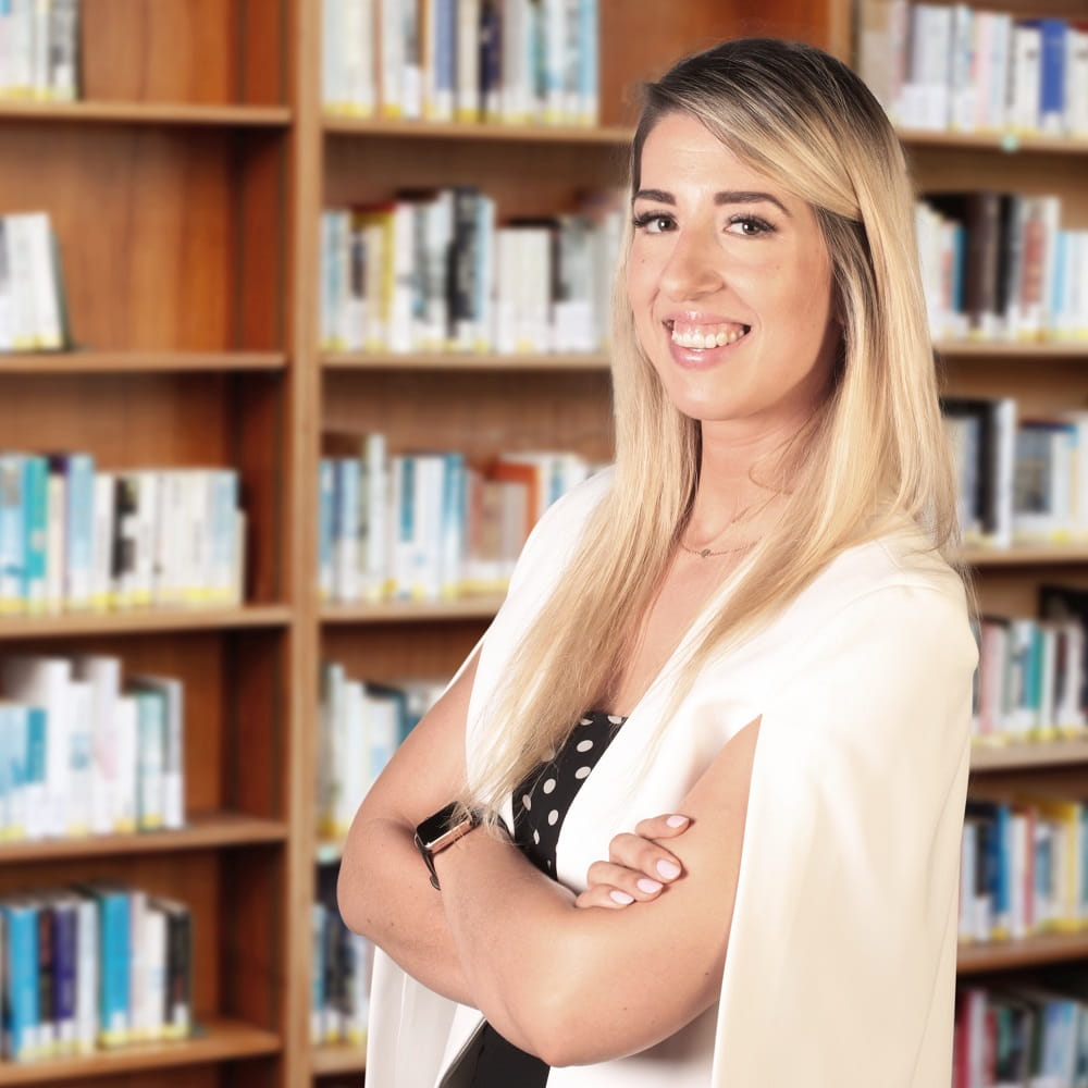 A blonde woman wearing a white top, standing in front of some bookcases with her arms folded across her chest, smiling at the camera.