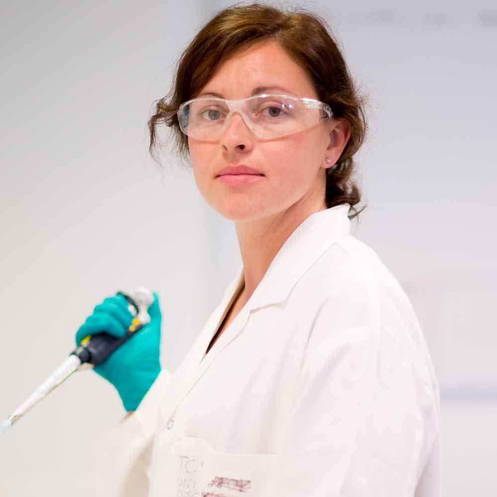 A female academic, wearing a white lab coat and plastic goggles, holds a piece of scientific equipment in her hands 