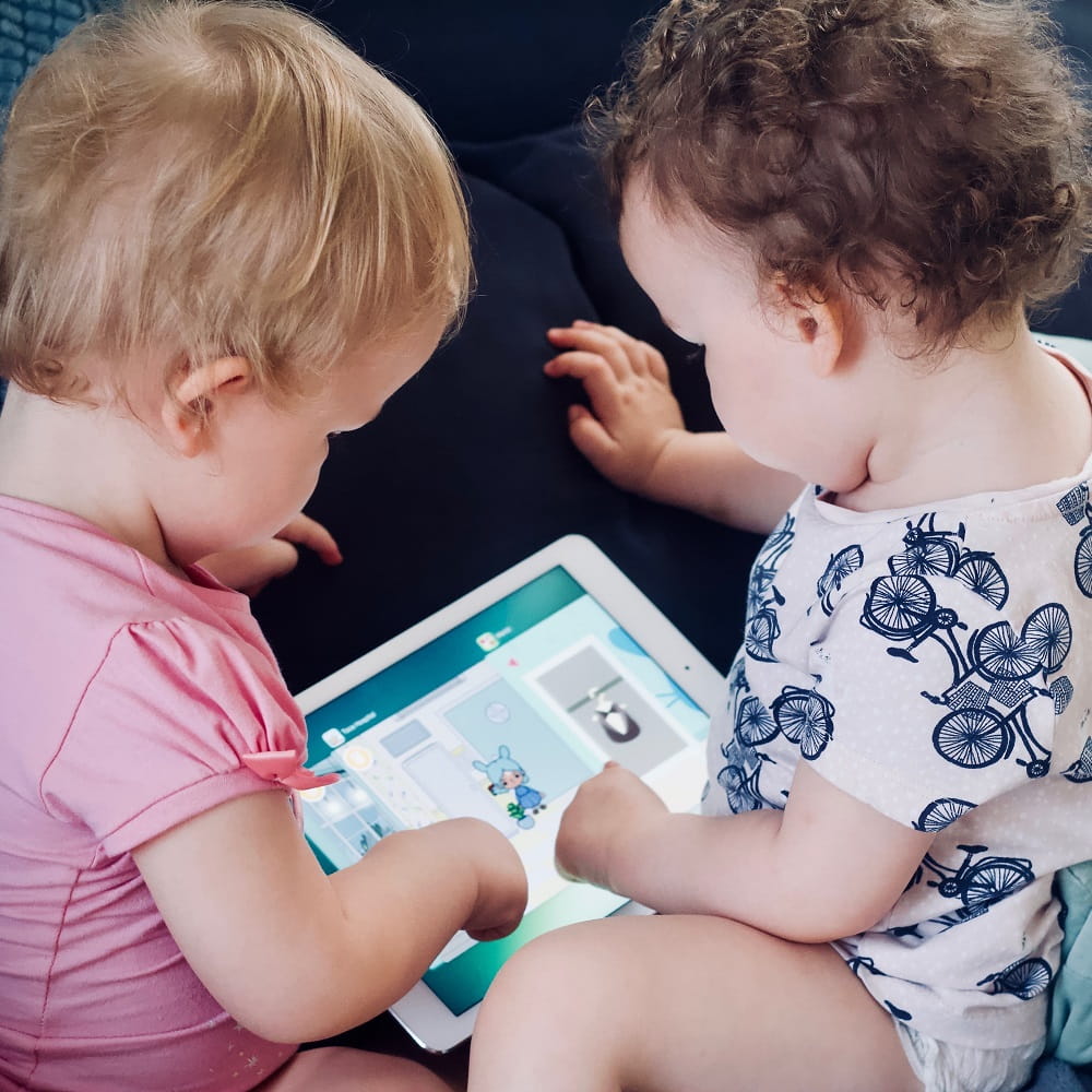 Two babies, sitting with their backs to the camera, looking at an iPad that is lying between them.