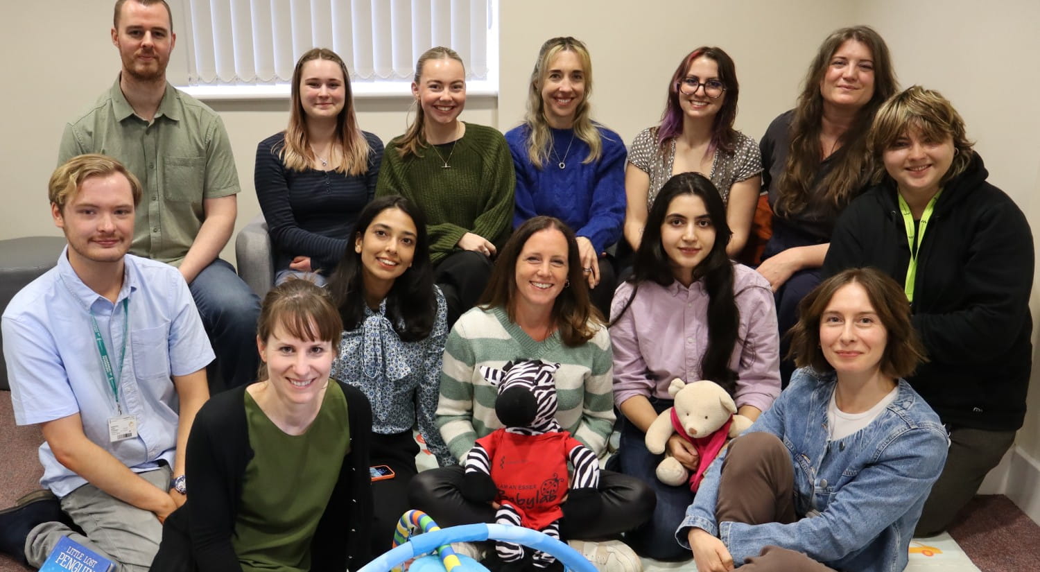A photo of 13 men and women, the researchers from the Baby Lab, sitting in a room and smiling at the camera.