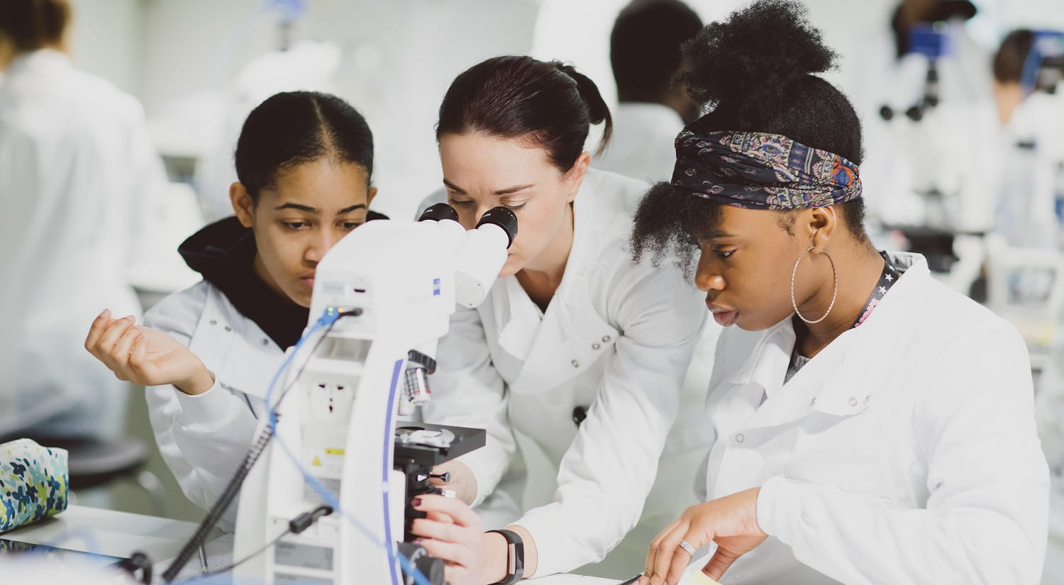 A lecturer looking down a digital microscope, a female student standing on either side of her.