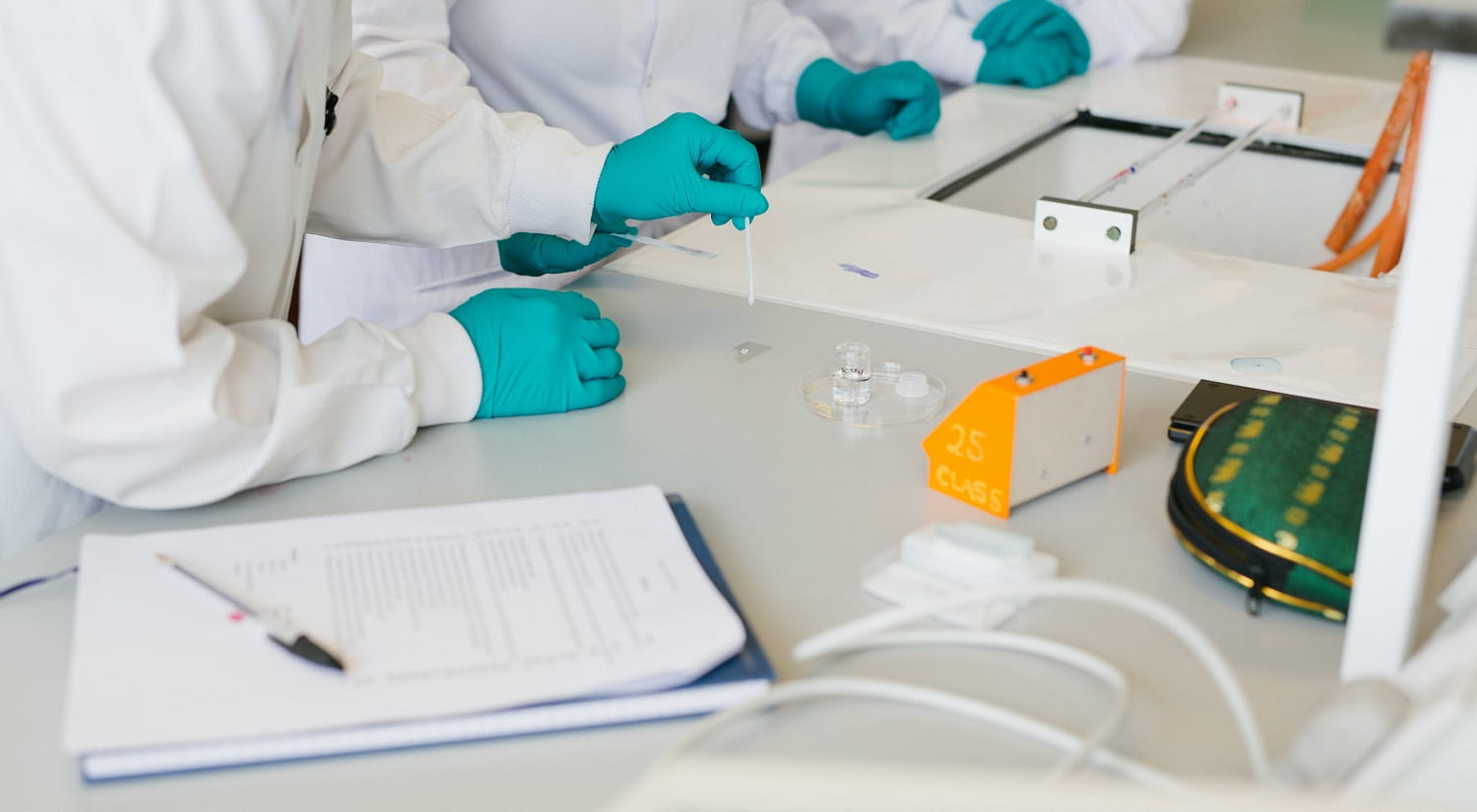 Three people in white coats wearing teal gloves, one of them is using a cotton bud to add something to a microscope slide sitting on a lab bench. A notepad and some paper with a pen on top are in the foreground.