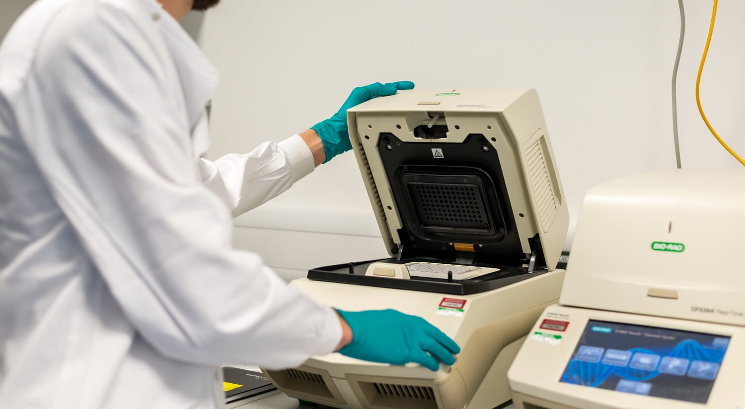 A person in a white lab coat and green gloves, lifting the lid on a piece of life sciences laboratory equipment.